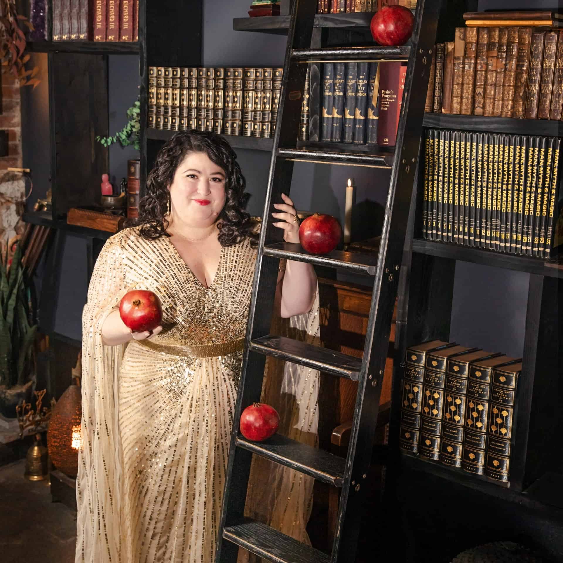 Kara Loewentheil holding a pomegranate in front of a bookshelf in a gold dress.