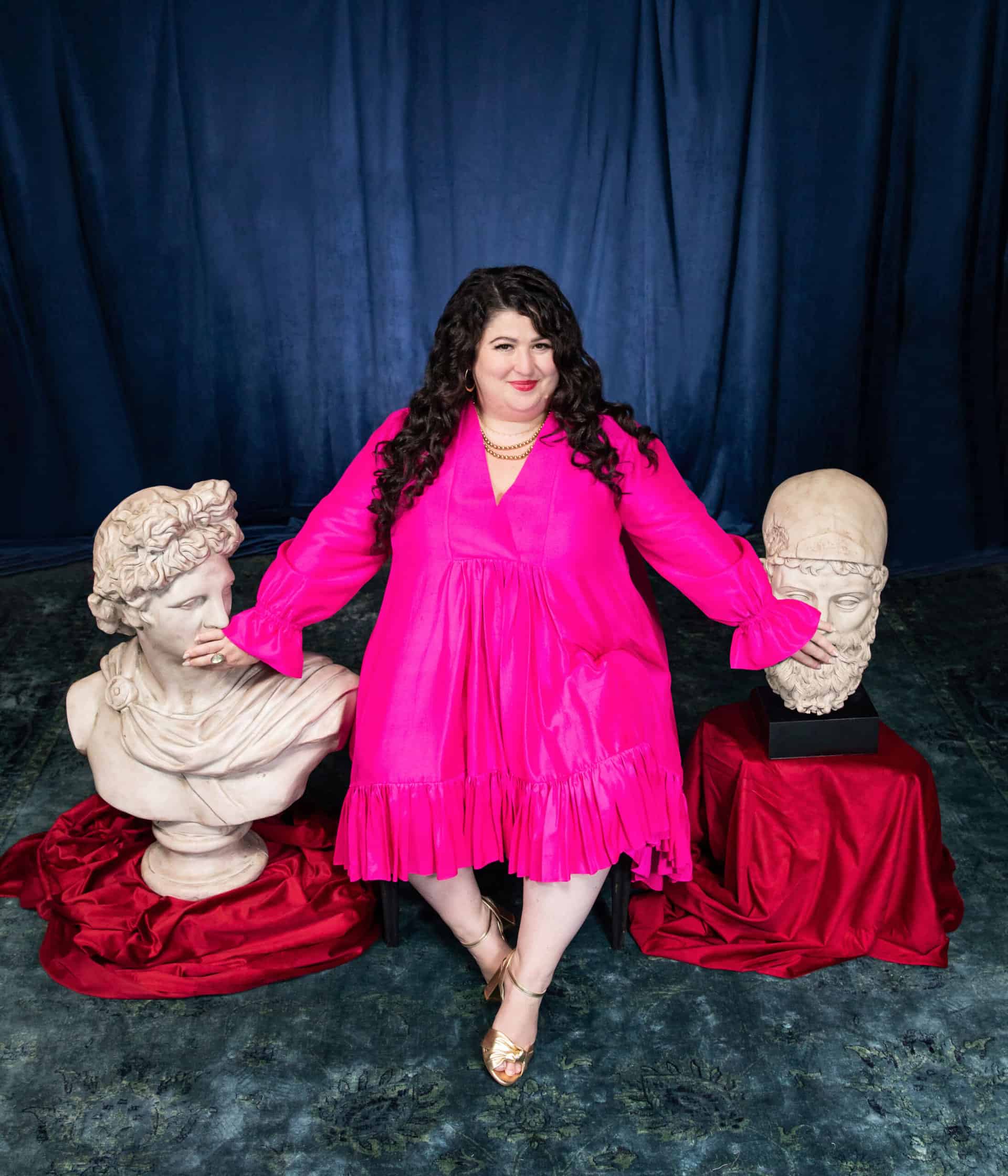 Kara Loewentheil sitting between two sculpture busts in a pink dress.